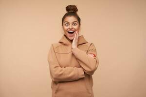 Excited young pretty brunette female dressed in nude sweatshirt holding palm on her cheek while looking amazedly at camera with wide eyes and mouth opened, isolated over beige background photo