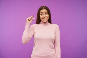 Horizontal shot of young lovely brown haired woman biting her underlip while looking ironically on raised hand and showing small size with her fingers, posing over purple background photo