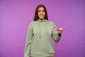 Bewildered young lovely brown haired female biting worringly underlip while showing aside with forefinger, isolated over purple background in casual clothes photo