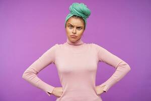 Studio photo of young serious brown haired woman with natural makeup frowning her eyebrows while looking crossly at camera, isolated over purple background