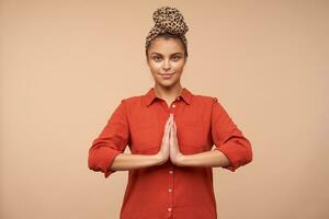 Positive young green-eyed brunette elegant lady with natural makeup looking calmly at camera and keeping raised hands together, isolated over beige background photo