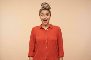 Dazed young elegant brown haired woman wearing her headband in knot while posing over beige background, looking amazedly at camera with wide eyes and mouth opened photo