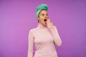 Tired young elegant brunette female with red manicure keeping her eyes closed while yawning and closing her mouth with raised palm, standing over purple background photo