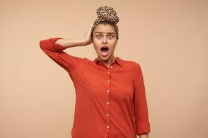 Bewildered young pretty brown haired lady rounding surprisedly her green eyes and keeping raised hand on her head, isolated over beige background in casual wear photo