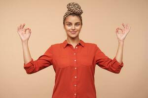 Positive young pretty brown haired woman with headband smiling slightly while looking at camera and folding fingers in mudra gesture, isolated over beige background photo