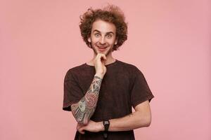 Joyful young attractive curly redhead male with tattooes looking excitedly at camera and holding his chin with raised hand, wearing dark red t-shirt while standing over pink background photo