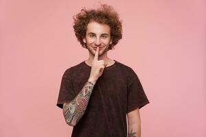 Indoor photo of positive young curly bearded tattooed male raising forefinger to his mouth and smiling pleasantly, asking to keep secret, isolated over pink background