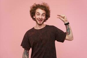 Excited young pretty curly bearded tattooed guy raising his eyebrows while looking to camera and showing small size with his fingers, dressed in dark red t-shirt while posing over pink background photo