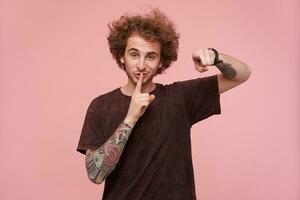 Shot of young pretty curly bearded redhead guy with tattooes raising hand in hush gesture and pointing at camera with forefinger, wearing dark red t-shirt over pink background photo