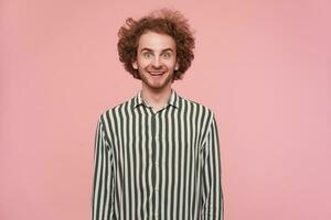 Surprised young lovely redhead male with beard rounding his green eyes while looking amazedly at camera and smiling cheerfully, isolated over pink background in casual wear photo