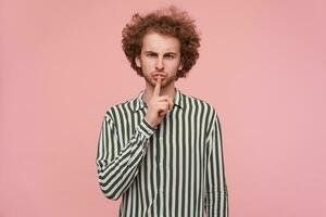 Studio photo of serious young bearded redhead male raising hand in hush gesture while posing over pink background, frowning his eyebrows while asking to keep silence