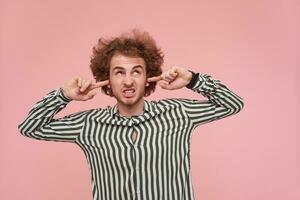 Displeased young bearded curly male with casual hairstyle inserting fingers into his ears while trying to avoid loud sounds, frowning face while posing over pink background photo