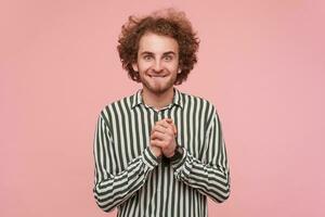 emocionado joven ojos verdes barbado pelirrojo hombre vestido en casual ropa plegable elevado manos y mordiendo debajo del labio mientras en pie terminado rosado antecedentes en a rayas camisa foto