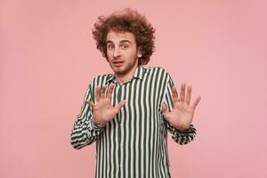 Portrait of attractive young curly redhead guy with beard raising palms in protective gesture and looking scaredly at camera, wearing striped shirt while standing over pink background photo