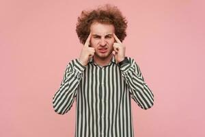 Unhappy young bearded curly redhead guy keeping forefingers on his temple and frowning his face while having strong headache, standing over pink background photo