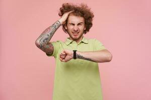 Indoor photo of young curly brunette man with tattoos grimacing his face while checking time with his wristwatch, standing over pink background in green polo