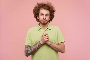 Indoor shot of young brunette guy with tattoos folding hands in praying gesture and looking desperately at camera, standing over pink background in casual wear photo