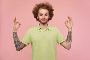 Good looking young curly brown haired tattooed man keeping his eyes closed while meditating and folding fingers in mudra gesture, isolated over pink background photo