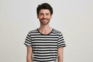 Portrait of cheerful attractive young man with bristle wears striped t shirt feels happy, standing and smiling isolated over white background Looks directly in camera photo