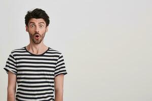 Closeup of surprised handsome young man with bristle wears striped t shirt feels stunned standing over white wall with copyspace for your advertising photo