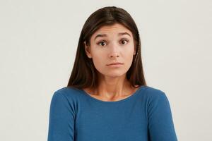 Closeup of amazed pretty young woman with long hair wears navy longsleeve feels embarrassed and sad standing over white wall with copyspace for your advertising photo