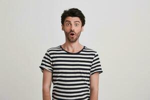 Portrait of amazed attractive young man with bristle and opened mouth wears striped t shirt feels surprised and shocked isolated over white background Looks directly in camera photo