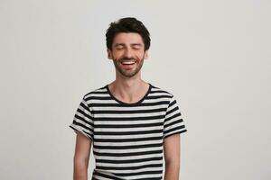 Closeup of smiling attractive young man with bristle wears striped t shirt keeps eyes closed and feels excited standing over white wall with copyspace for your advertising photo