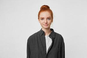 Nice red-haired girl with a bun on her head dressed in a simple oversize black shirt is looking directly at the camera with a slight gentle smile, isolated on white background photo