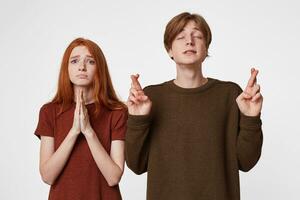 Guy with his eyes closed, fingers crossed, makes a wish, hopes for happy circumstances.Red-haired girl holds palms together,with a praying look, hoping the higher forces will help her photo
