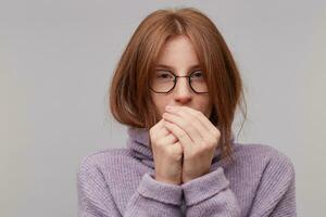 Young attractive red-haired girl in glasses dressed in a warm purple sweater with a high collar blowing on cold palms trying to get warm photo