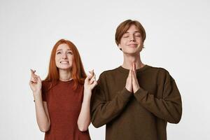 Red-haired girl on a white background is standing on a raised head and hands up crossed her fingers in hope of something. Guy stands with his eyes closed and pacified smile, folded his hands in prayer photo