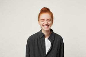 Cheerful resilient red-haired girl with a bun at her head laughs sincerely, having closed her eyes, dressed in a simple black shirt, is isolated on white background photo
