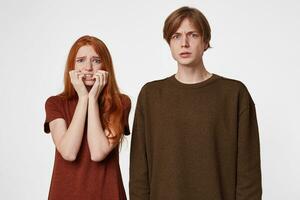 Portrait of scared couple, girl looks frightened afraid chattering teeth with fear,bites nails from fright, hands holding cheeks, the guy looks tense feeling threatened, over white background photo