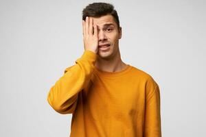 Portrait of irresponsible man having troubles. Frightened man in despair covered one eye with his palm, dressed casually, isolated over white background photo