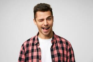 Grinning broadly handsome attractive charming young man, winks with one eye, flirting smile, pleasant appearance, dressed in checkered shirt, standing against white background. photo