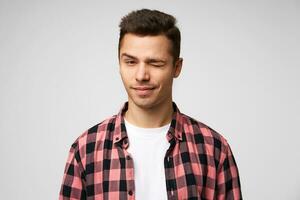 Close up of a flirting handsome attractive charming guy looking confidently,winks with one eye, slightly smiling, pleasant appearance, dressed in checkered shirt, standing against white background. photo