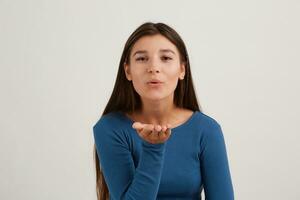 Cute looking woman, beautiful girl with dark long hair. Wearing blue jumper. Emotion concept. Blow over palm, sending air kiss. Watching at the camera isolated over white background photo