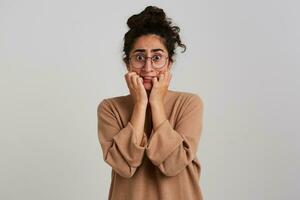 Portrait of horrified, adult girl with dark curly hair bun. Wearing beige jumper and glasses. Emotion concept. Touching her face in fear. Watching at the camera isolated over white background photo