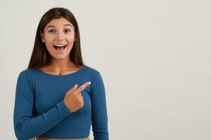 Happy looking woman, beautiful girl with dark long hair. Wearing blue jumper. Emotion concept. Watching at the camera and pointing finger to the right at copy space, isolated over white background photo