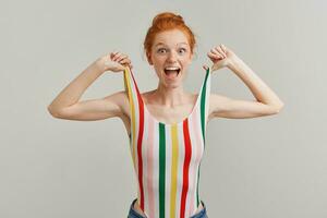 Positive looking woman, beautiful redhead girl with hair bun and freckles. Wearing striped colorful swimsuit. Pulls straps up. Emotion concept. Watching at the camera isolated over grey background photo