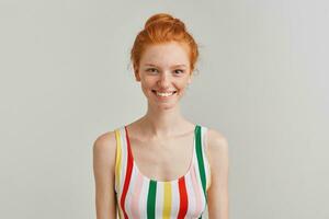 Cheerful lady, positive woman with ginger hair bun and freckles. Wearing striped colorful swimsuit. Emotion concept. Watching at the camera and smiling isolated over grey background photo