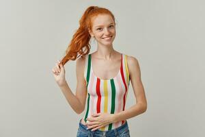 Portrait of attractive, happy girl with ginger pony tail and freckles. Wearing striped colorful swimsuit and jeans. Holding strand of hair. Watching at the camera isolated over grey background photo