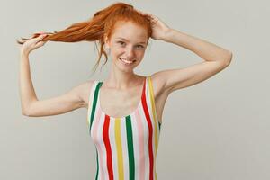 Cute woman, beautiful redhead girl with pony tail and freckles. Wearing striped colorful swimsuit. Touching her head and holding strand of hair. Watching at the camera isolated over grey background photo