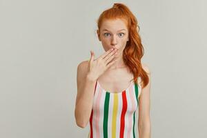 Portrait of shocked, surprised girl with ginger pony tail and freckles. Wearing striped colorful swimsuit. Emotion concept. Keeps palm over mouth. Watching at the camera isolated over grey background photo