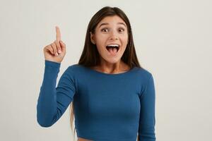 Portrait of attractive, excited girl with dark long hair. Wearing blue jumper. Emotion concept. Raises finger up, got an idea. Watching at the camera isolated over white background photo