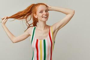 Positive lady, pretty woman with ginger pony tail and freckles. Wearing striped colorful swimsuit. Touching her head and holding strand of hair. Watching at the camera isolated over grey background photo