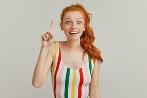 Portrait of amazed girl with ginger pony tail and freckles. Wearing striped colorful swimsuit. Emotion concept. Raises finger up, got an idea. Watching at the camera isolated over grey background photo