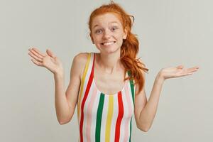 Young lady, pretty woman with ginger pony tail and freckles. Wearing striped colorful swimsuit. Emotion concept. Shrugs her shoulders and smile. Watching at the camera isolated over grey background photo