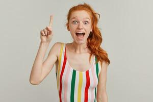 Surprised lady, amazed woman with ginger pony tail and freckles. Wearing striped colorful swimsuit. Emotion concept. Raises finger up, got an idea. Watching at the camera isolated over grey background photo