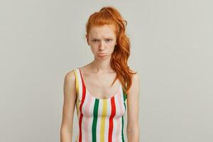 Discontent lady, sad woman with ginger pony tail and freckles. Wearing striped colorful swimsuit. Emotion concept. Frowns and pouts her lip. Watching at the camera isolated over grey background photo